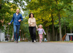 Familie mit Kind läuft fröhlich auf einem Campingplatzweg, umgeben von Bäumen.
