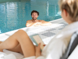 Mann entspannt im Pool und lächelt Frau an, die im Vordergrund auf ihr Handy schaut.