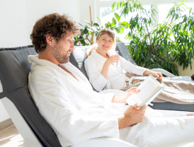 Ruhebereich vom Baltic Spa liest ein Mann im Bademantel ein Buch, während eine Frau neben ihm entspannt und ein Glas Wasser hält.