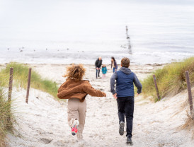 Zwei Kinder rennen freudig durch die Dünen zum Strand, während eine Familie im Hintergrund am Meer steht.