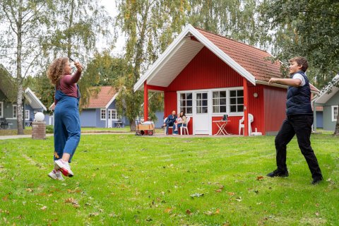 Ein Mädchen und ein Junge spielen Badminton im Vordergrund, im Hintergrund Familie vor einer roten Hütte im Campingplatz Baltic Freizeit in Markgrafenheide.