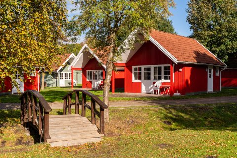 Rote und grüne Ferienhäuser umgeben von Bäumen auf dem Campingplatz Baltic Freizeit in Markgrafenheide mit einer Holzbrücke im Vordergrund.