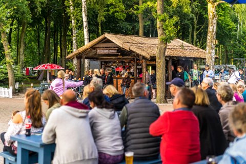 Gemütliche Outdoor-Bar im Wald mit vielen Gästen und entspannter Sommerstimmung.          ChatGPT fragen
