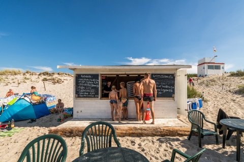 Weiße Strandbar im Sand mit Getränken, Eis und Badegästen bei Sonnenschein.          ChatGPT fragen