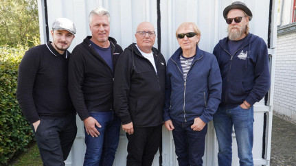 Fünf Bandmitglieder stehen lässig nebeneinander vor einem rostigen Metallcontainer. Sie tragen Sonnenbrillen, T-Shirts, Jeans und Sneakers – der Look: authentisch, cool und bodenständig. Die Haltung und der Gesichtsausdruck strahlen Selbstbewusstsein und Zusammenhalt aus – eine echte Rockband mit Charakter.