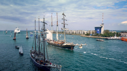 Mehrmastige Traditionssegler und moderne Schiffe kreuzen bei strahlendem Wetter durch die Warnemünder Hafeneinfahrt. Im Vordergrund fahren zwei imposante Segelschiffe mit zahlreichen Personen an Bord, während im Hintergrund Kreuzfahrtschiffe, Yachten und ein markanter blauer Hafenturm das maritime Flair der Hanse Sail unterstreichen. Die Szene vermittelt lebendiges Treiben und festliche Stimmung auf dem Wasser.