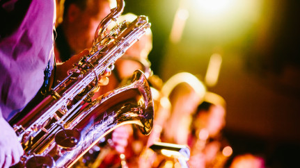 Nahaufnahme einer Jazz-Performance: Im Vordergrund glänzt ein Saxophon im warmen Bühnenlicht, die Musiker:innen im Hintergrund sind unscharf, aber sichtbar vertieft ins Spiel. Die Szene ist in leuchtenden Gold- und Violetttönen gehalten und vermittelt die energiegeladene, stimmungsvolle Atmosphäre eines Live-Konzerts.