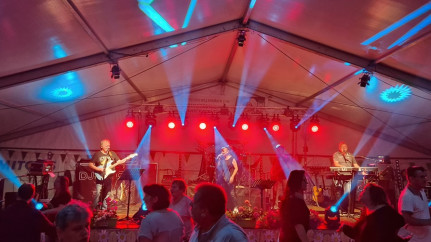 Livemusik im Festzelt: Die Band „The Liberty’s“ steht auf einer hell ausgeleuchteten Bühne, umgeben von roten und blauen Lichtstrahlen. Gitarrist, Sängerin und Keyboarder performen vor einem tanzenden Publikum, das sich sichtbar von der mitreißenden Atmosphäre anstecken lässt. Die Stimmung ist lebendig, sommerlich und festlich.