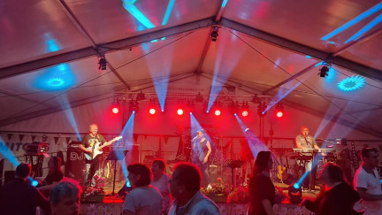 Livemusik im Festzelt: Die Band „The Liberty’s“ steht auf einer hell ausgeleuchteten Bühne, umgeben von roten und blauen Lichtstrahlen. Gitarrist, Sängerin und Keyboarder performen vor einem tanzenden Publikum, das sich sichtbar von der mitreißenden Atmosphäre anstecken lässt. Die Stimmung ist lebendig, sommerlich und festlich.