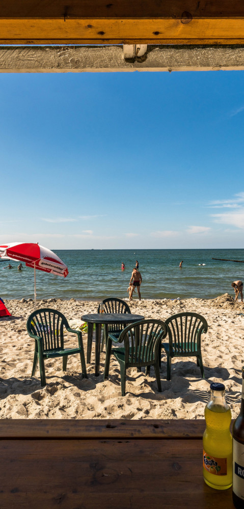 Blick vom Innenbereich eines Holzpavillons auf den Sandstrand mit Sonnenschirmen, Zelten, Badegästen und Erfrischungsgetränken am Ufer der Ostsee.