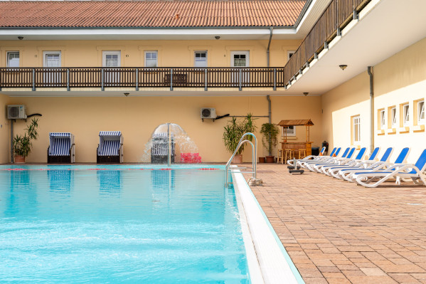 Schwimmbad mit Liegestühlen und Brunnen am Campingplatz, umgeben von Gebäuden.