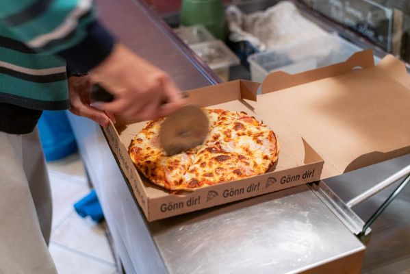 Mitarbeiter schneidet frisch gebackene Pizza in einer Kartonbox mit dem Text 'Gönn dir!' am Campingplatz.