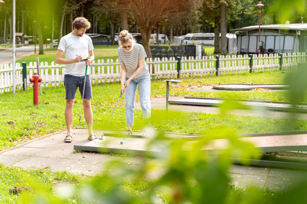 Junges Paar spielt Minigolf auf dem Gelände von Baltic Freizeit, umgeben von grüner Natur und Wohnmobilen."