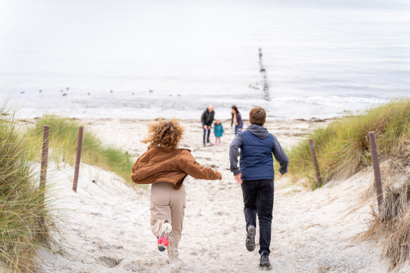 Zwei Kinder rennen freudig durch die Dünen zum Strand, während eine Familie im Hintergrund am Meer steht.