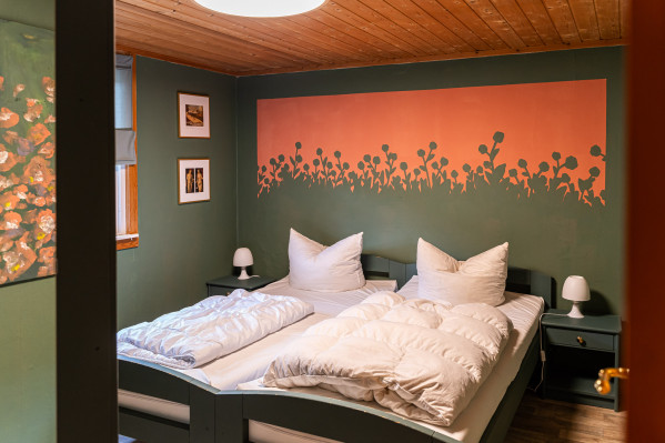 Schlafzimmer mit Doppelbett, grünen Wänden, orangefarbenem Blumenwandbild und Holzdecke neben einer Fensterseite.