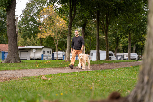 Camper geht mit seinem Hund durch den Campingplatz von Baltic Freizeit umgeben von Bäumen und Wohnwagen.