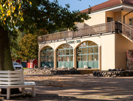 Baltic-SPA-Gebäude auf einem Campingplatz, flankiert von Bäumen mit herbstlichen Blättern und einer Bank im Vordergrund.