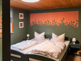 Schlafzimmer mit Doppelbett, grünen Wänden, orangefarbenem Blumenwandbild und Holzdecke neben einer Fensterseite.