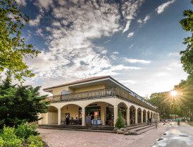 Gebäude mit Pool-Bereich, Restaurant und Wellness-Bereich auf dem Campingplatz von Baltic Freizeit in Markgrafenheide bei Sonnenuntergang mit Besuchern und umgebenden Bäumen.