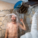 Mann genießt kalte Dusche aus einem Holzeimer in rustikaler Umgebung.