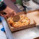 Mitarbeiter schneidet frisch gebackene Pizza in einer Kartonbox mit dem Text 'Gönn dir!' am Campingplatz.