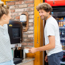 Mann holt sich Kaffee am Kaffeeautomat, während er sich lächelnd mit einer Frau unterhält.