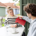 Lachende Frau in gestreiftem Pullover empfängt Geschenktüte von Mann auf einer Terrasse mit Campinghütten im Hintergrund.