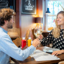 Frau und Mann sitzen sich lächelnd gegenüber am Tisch und stoßen mit einem Glas Wein im Restaurant auf dem Campingplatz an.